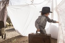 Elementary age boy wearing hat carrying luggage in outdoor tent made of sheets — Stock Photo
