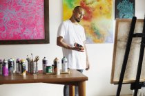Artista japonés de pie en la galería de arte, sosteniendo lata de pintura en aerosol, mirando obras de arte en caballete . - foto de stock
