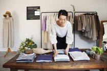 Japanese woman standing in a small fashion boutique, arranging t-shirts on a coffee table. — Stock Photo