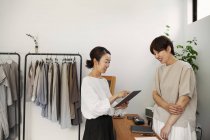 Две улыбающиеся японки стоят в маленьком модном бутике, держа в руках цифровой планшет . — стоковое фото