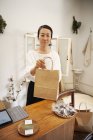 Sonriente mujer japonesa de pie en una pequeña boutique de moda, sosteniendo bolsa de compras de papel marrón, mirando en cámara
. - foto de stock