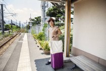 Japanese woman wearing hat standing on railway station platform with shopping bag and pink suitcase. — стокове фото