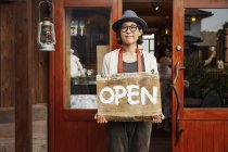 Femme japonaise portant chapeau et lunettes debout devant une boutique de cuir, tenant signe ouvert . — Photo de stock