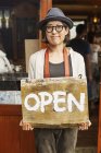 Japanese woman wearing hat and glasses standing in front of a leather shop, holding Open sign. — Stock Photo