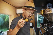 Japanese man wearing hat and glasses standing in an Eco Cafe, smelling freshly roasted coffee beans. — Stock Photo