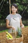 Sorrindo mulher japonesa vestindo avental em pé ao ar livre, segurando cesta com frutas e legumes frescos, olhando na câmera . — Fotografia de Stock