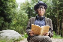 Японська жінка в окулярах і капелюсі сидить на стільці біля Еко Кафе, читаючи книжку. — стокове фото