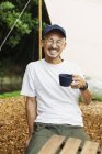 Homem japonês sorrindo vestindo boné de beisebol e óculos sentados ao ar livre em uma mesa, bebendo xícara de café . — Fotografia de Stock