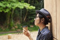 Japonesa mujer usando gafas y sombrero de pie fuera de Eco Café, sosteniendo taza de papel, vista lateral . - foto de stock
