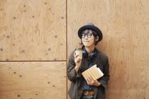 Mujer japonesa con gafas y sombrero de pie fuera de Eco Café, sosteniendo taza de papel y cuaderno . - foto de stock