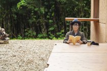 Femme japonaise portant des lunettes et un chapeau assis à une table à l'extérieur Eco Cafe, livre de lecture . — Photo de stock