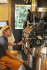 Japonais portant chapeau et lunettes assis dans un Eco Cafe, faisant fonctionner une machine de torréfaction de café . — Photo de stock