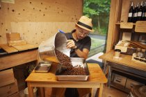 Japaner mit Hut und Brille sitzt in einem Öko-Café und gießt frisch geröstete Kaffeebohnen in Metalltablett. — Stockfoto