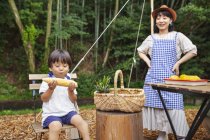 Japanese woman standing outdoors, wearing hat and apron and boy sitting on chair, eating corn on the cob. — Stock Photo