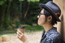 Japanese woman in glasses and hat standing outside Eco Cafe, holding paper cup. — Stock Photo