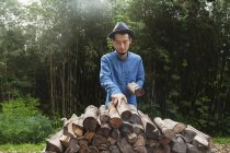 Japanese man wearing hat standing outdoors, stacking logs of firewood. — Stock Photo