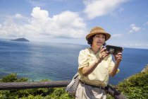Japanese woman wearing hat standing on a cliff, taking selfie with mobile phone. — Stock Photo