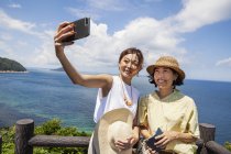 Две японки в шляпах, стоящих на утесе, делают селфи с помощью мобильного телефона. . — стоковое фото