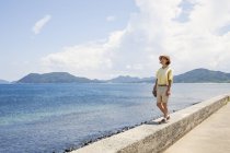 Japanese woman wearing hat standing on a wall, looking at ocean. — Stock Photo