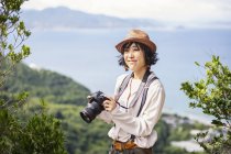 Japanese woman carrying backpack and holding camera standing on a cliff by ocean. — Stock Photo