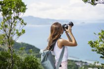 Japanese woman carrying backpack taking photograph on a cliff by ocean. — Stock Photo