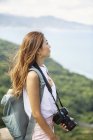 Japanese woman carrying backpack and holding camera standing on a cliff by ocean. — Stock Photo