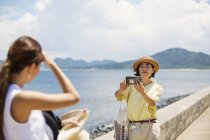 Two Japanese women standing by the ocean, taking picture with mobile phone. — Stock Photo