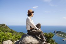 Japanerin mit Hut sitzt auf einem Felsen an einer Klippe mit Meereslandschaft. — Stockfoto