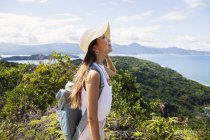 Japonesa mujer usando sombrero y llevando mochila de pie en un acantilado con paisaje oceánico . - foto de stock