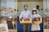 Japanese man and woman standing outside a farm shop, holding crate and basket with fresh vegetables, looking in camera. — Stock Photo