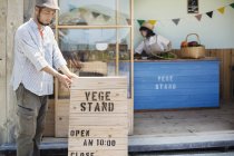 Japanese man wearing cap standing outside a farm shop, putting up wooden shop sign. — Stock Photo