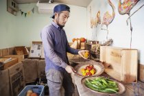 Japanese man wearing cap standing in farm shop, holding bowls with fresh vegetables. — Stock Photo