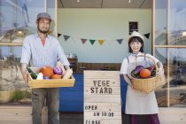 Japonés hombre y mujer de pie fuera de una tienda de granja, sosteniendo cajón y cesta con verduras frescas, mirando en la cámara
. - foto de stock