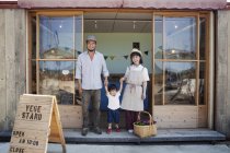 Japonés hombre, mujer y niño de pie fuera de una tienda de granja, tomados de la mano, mirando en la cámara
. - foto de stock