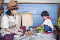 Femme japonaise portant chapeau et garçon assis devant un magasin de ferme, plantant des fleurs dans des pots de fleurs . — Photo de stock