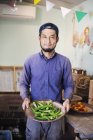 Smiling Japanese man wearing cap standing in farm shop, holding bowl with fresh okra. — Stock Photo
