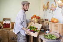 Smiling Japanese man wearing cap standing in farm shop, holding crate with fresh vegetables. — Stock Photo