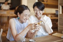 Due donne giapponesi sedute a un tavolo in un caffè vegetariano, utilizzando il telefono cellulare
. — Foto stock