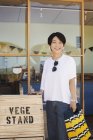 Femme japonaise debout devant un magasin de ferme, tenant un sac à provisions, souriant à la caméra . — Photo de stock