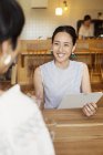 Smiling Japanese woman sitting with friend at a table in a vegetarian cafe, holding menu. — Stock Photo
