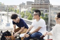 Drei junge japanische Männer sitzen auf einem Dach in urbaner Umgebung und trinken Bier. — Stockfoto