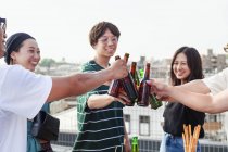 Grupo de jóvenes japoneses hombres y mujeres de pie en la azotea en el entorno urbano, tostando botellas de cerveza . - foto de stock