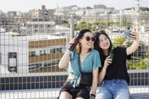 Due giovani donne giapponesi sedute sul tetto in ambiente urbano, scattano selfie con il cellulare e tengono in mano bottiglie di birra . — Foto stock