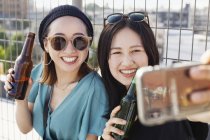 Zwei junge Japanerinnen sitzen auf einem Hausdach in urbaner Umgebung, machen ein Selfie mit dem Handy und halten Bierflaschen in der Hand. — Stockfoto