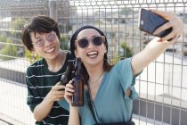 Young Japanese man and woman sitting on rooftop in urban setting, taking selfie with mobile phone. — Stock Photo