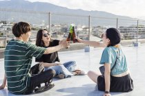 Jovem japonês homem e mulheres sentados no telhado em ambiente urbano, brindando com garrafas de cerveja . — Fotografia de Stock