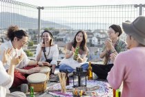 Группа молодых японских мужчин и женщин, сидящих на крыше в городских условиях, пьющих пиво и играющих на барабанах . — стоковое фото