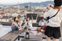 Smiling group of young Japanese man and women on rooftop in urban setting, drinking beer and posing as woman taking photo. — Stock Photo