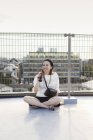 Sorridente giovane donna giapponese seduta sul tetto in ambiente urbano, bere birra . — Foto stock