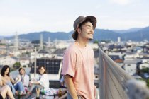 Sonriente joven japonés de pie en la azotea en el entorno urbano, amigos de fondo
. - foto de stock